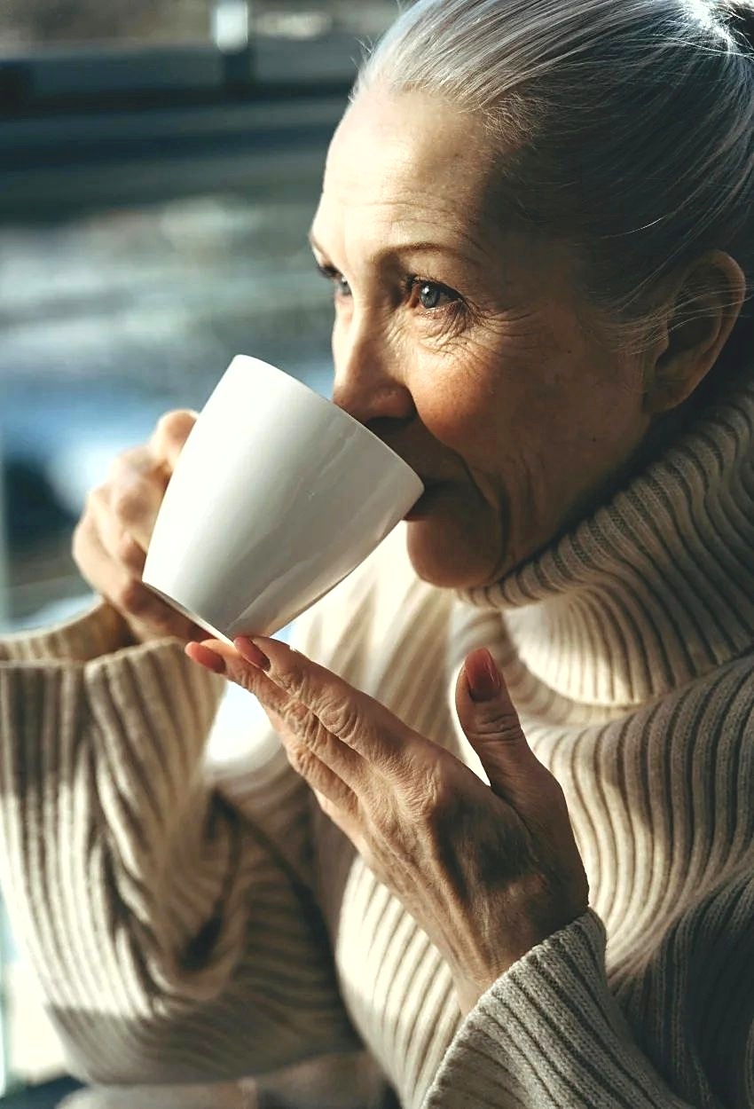 A woman in her seventies drinking coffee by a window in daylight, looking out.